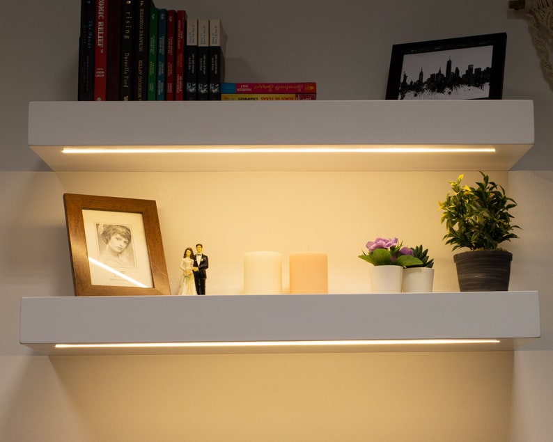 May include: Two white floating shelves with built-in LED lighting. The top shelf holds books and a framed cityscape print. The bottom shelf displays a framed photo, candles, small plants, and a wedding cake topper. The shelves are mounted on a neutral-colored wall.