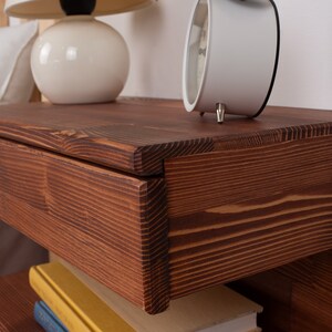 May include: A wall-mounted floating nightstand crafted from dark stained wood with a prominent grain pattern. The rectangular unit features a single drawer with a clean front edge and an open storage shelf underneath for books or other small items.