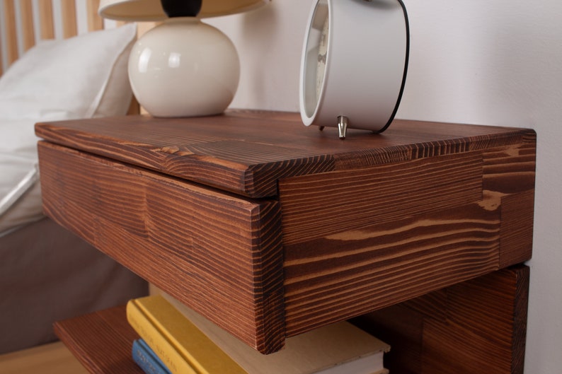 May include: A wall-mounted floating nightstand crafted from dark stained wood with a prominent grain pattern. The rectangular unit features a single drawer with a clean front edge and an open storage shelf underneath for books or other small items.