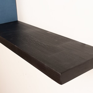 May include: A black wooden shelf with a natural wood grain finish. The shelf is mounted to a white wall and has a slight curve to it.