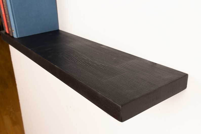 May include: A black wooden shelf with a natural wood grain finish. The shelf is mounted to a white wall and has a slight curve to it.
