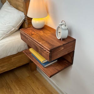 May include: A floating wooden nightstand with a dark brown wood grain finish, featuring a top surface holding a white lamp and a white analog alarm clock, and an open lower shelf containing two stacked books with yellow and blue covers.