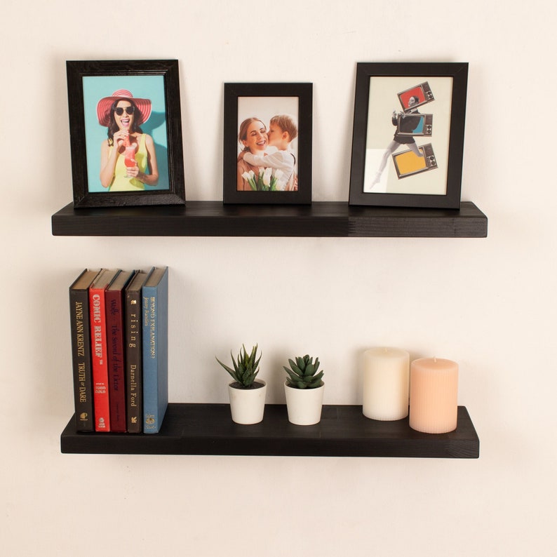 May include: Two black floating shelves with a variety of items on them, including framed photos, books, candles, and small plants. The shelves are mounted on a white wall.