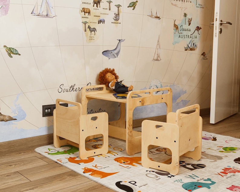 Puede incluir: Un conjunto de mesa y sillas de madera para ni&ntilde;os con un mapa del mundo en la pared detr&aacute;s. La mesa tiene un estante debajo y las sillas tienen respaldos abiertos. El conjunto est&aacute; sobre una alfombra con un patr&oacute;n de alfabeto.