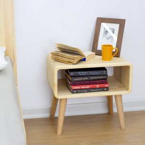 May include: A light wood nightstand with a rounded shelf and four legs. The nightstand has a book and a mug on top, and a stack of books on the shelf. The nightstand is next to a bed with a white pillow.