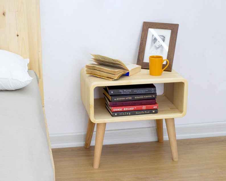 May include: A light wood nightstand with a rounded shelf and four legs. The nightstand has a book and a mug on top, and a stack of books on the shelf. The nightstand is next to a bed with a white pillow.