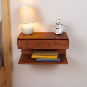 May include: A wall-mounted floating nightstand crafted from dark brown wood with a horizontal drawer and an open lower shelf. The top surface holds a white table lamp and a round white alarm clock, while the lower shelf holds two stacked books.