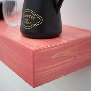 May include: A pink wooden shelf with a black watering can that says "Flowers & Garden" on it.