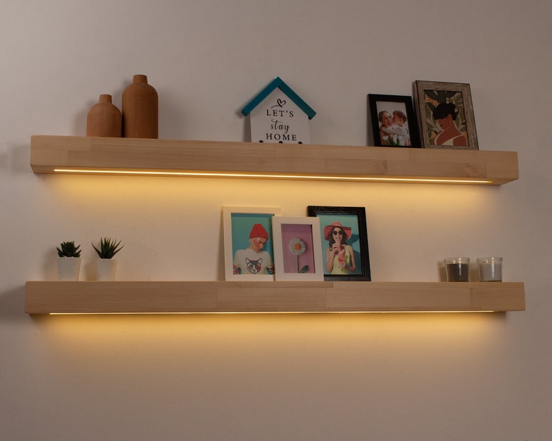 May include: Two wooden floating shelves with integrated LED lighting are mounted on a white wall. The top shelf holds two brown vases, a small house-shaped decoration, and framed photos. The bottom shelf displays framed photos, small potted plants, and candles.