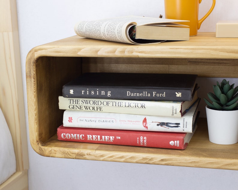 K&ouml;nnte beinhalten: Ein rundes Holzregal an der Wand mit einer Reihe von B&uuml;chern und einer kleinen Topfpflanze. Die B&uuml;cher tragen Titel wie "The Sword of the Lictor" und "Comic Relief".