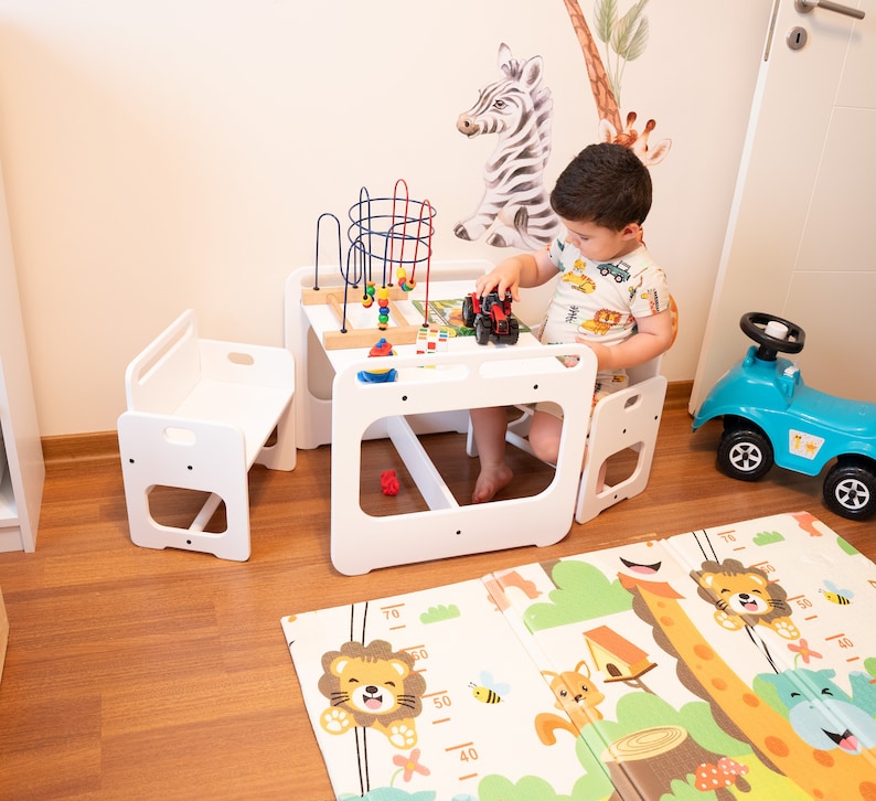 May include: A white wooden play table and two chairs with a colorful toy activity center on top. A child is sitting at the table playing with a toy truck. The table and chairs are on a wooden floor with a colorful play mat.