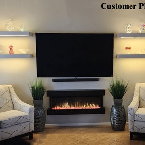 May include: A modern living room setup featuring a large black television mounted above a black electric fireplace. Two white armchairs with patterned upholstery flank the fireplace. Decorative shelves with illuminated displays are above the TV.