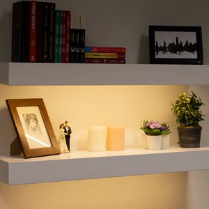 May include: Two white floating shelves with various decorative items. The top shelf holds books and a framed skyline print. The bottom shelf displays a framed photo, candles, small plants, and a figurine.