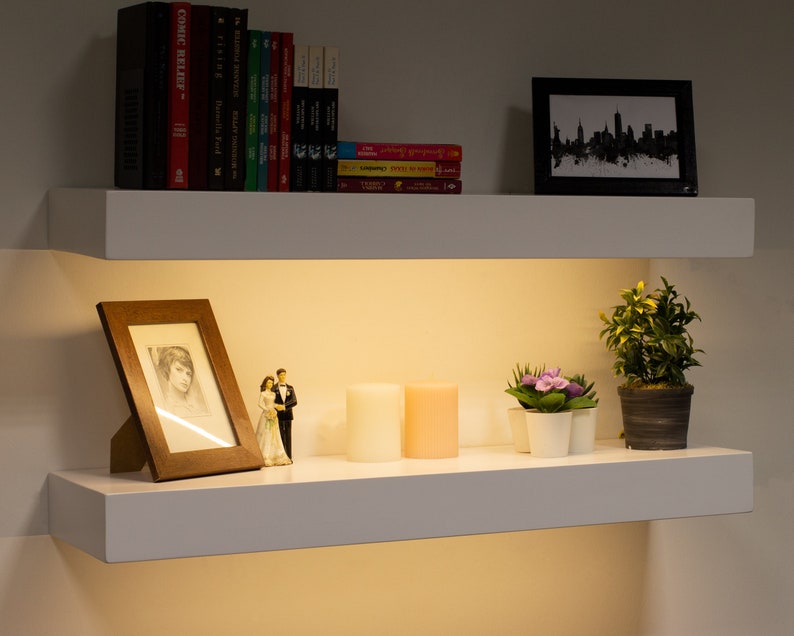 May include: Two white floating shelves with various decorative items. The top shelf holds books and a framed skyline print. The bottom shelf displays a framed photo, candles, small plants, and a figurine.