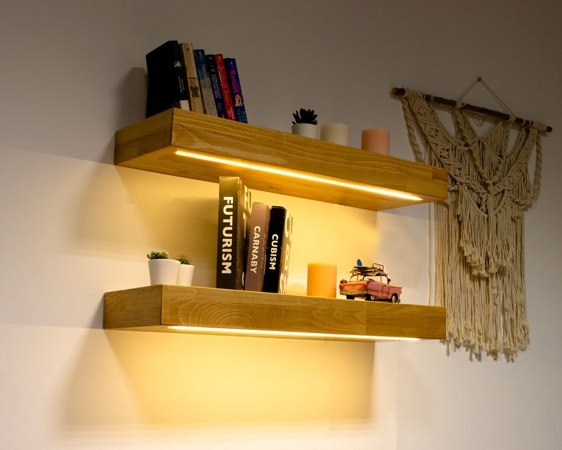 May include: Two wooden floating shelves with built-in LED lighting. The shelves are filled with books, small potted plants, candles, and a miniature red pickup truck. A macrame wall hanging is visible in the background.