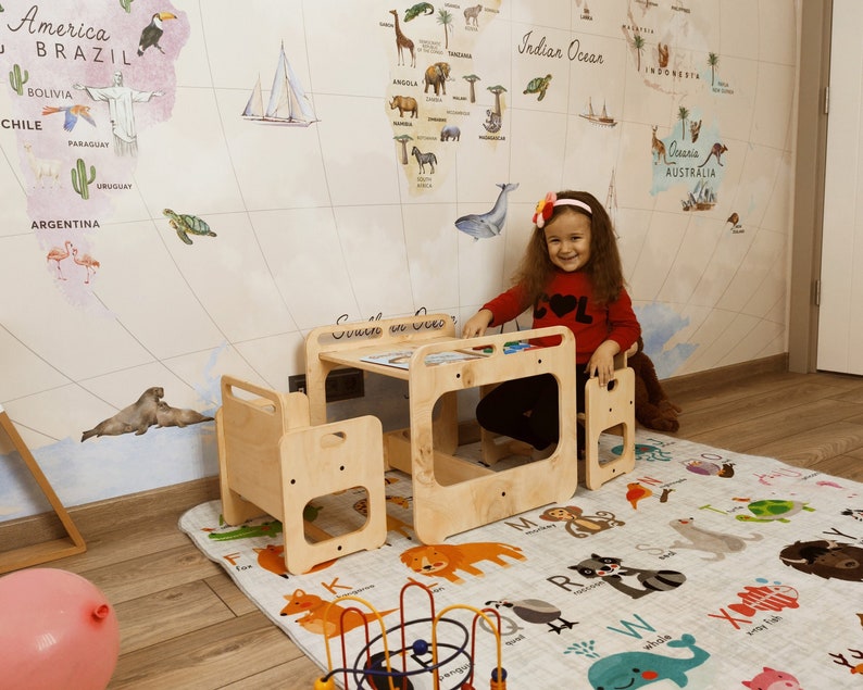 Puede incluir: Una ni&ntilde;a peque&ntilde;a se sienta en un conjunto de mesa y silla de madera para ni&ntilde;os. La mesa tiene un peque&ntilde;o estante debajo y la silla tiene un dise&ntilde;o recortado. El conjunto est&aacute; en una habitaci&oacute;n con un papel tapiz de mapa del mundo y una alfombra de alfabeto colorida.