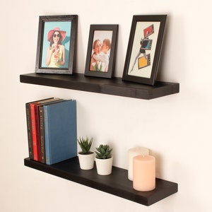 May include: Two black floating shelves with a variety of items on them, including framed photos, books, candles, and plants. The shelves are mounted on a white wall.