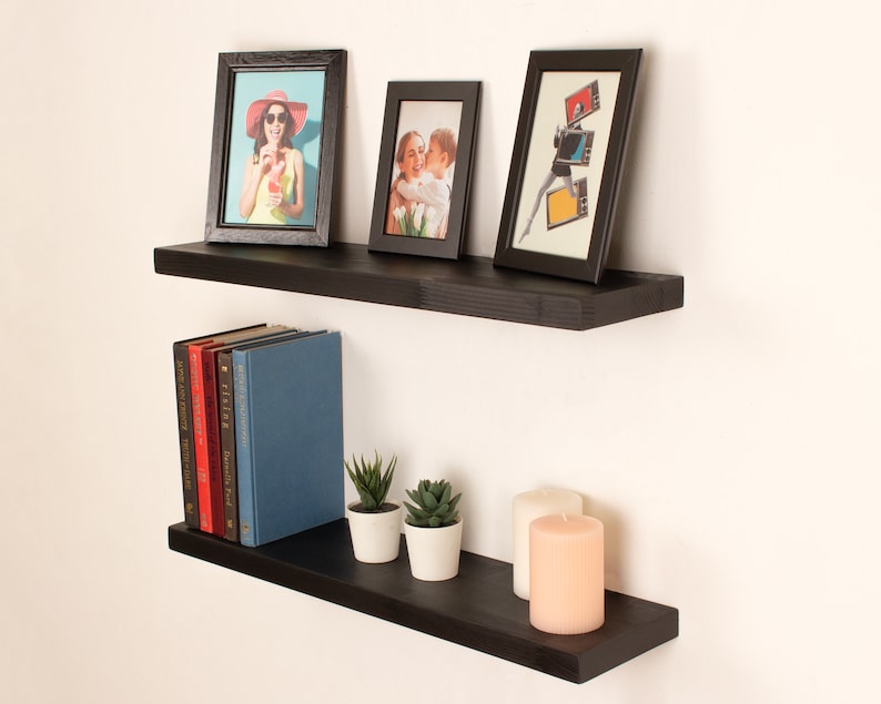 May include: Two black floating shelves with a variety of items on them, including framed photos, books, candles, and plants. The shelves are mounted on a white wall.