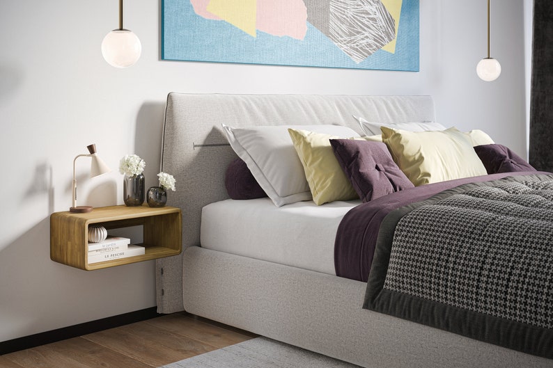 K&ouml;nnte beinhalten: Ein wei&szlig;es Bett mit einem grauen Kopfteil und einer grau-schwarzen gemusterten Decke. Das Bett hat einen schwebenden Nachttisch aus Holz mit zwei B&uuml;chern und einer Vase mit wei&szlig;en Blumen darauf. Auf dem Nachttisch steht eine Lampe und &uuml;ber dem Bett h&auml;ngt eine wei&szlig;e Kugel-Pendelleuchte.