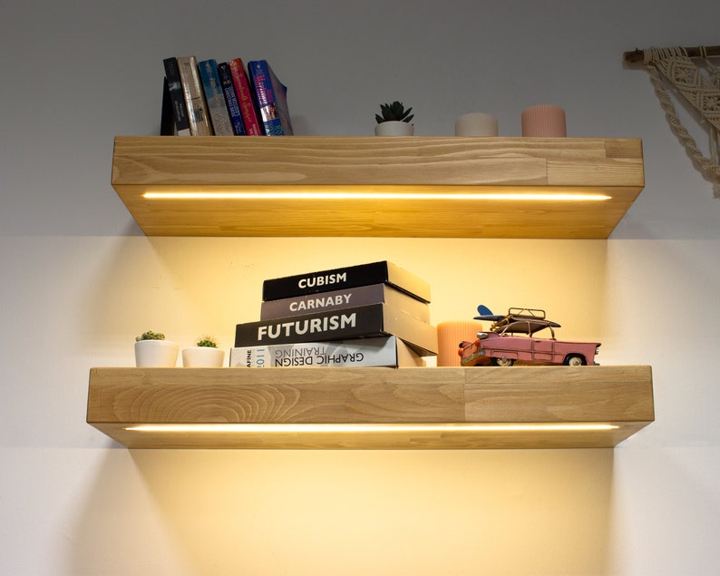May include: Two wooden floating shelves with built-in lighting are mounted on a white wall. The top shelf holds books and a small plant. The bottom shelf displays books with the titles "Cubism", "Carnaby", and "Futurism", along with a pink toy car.