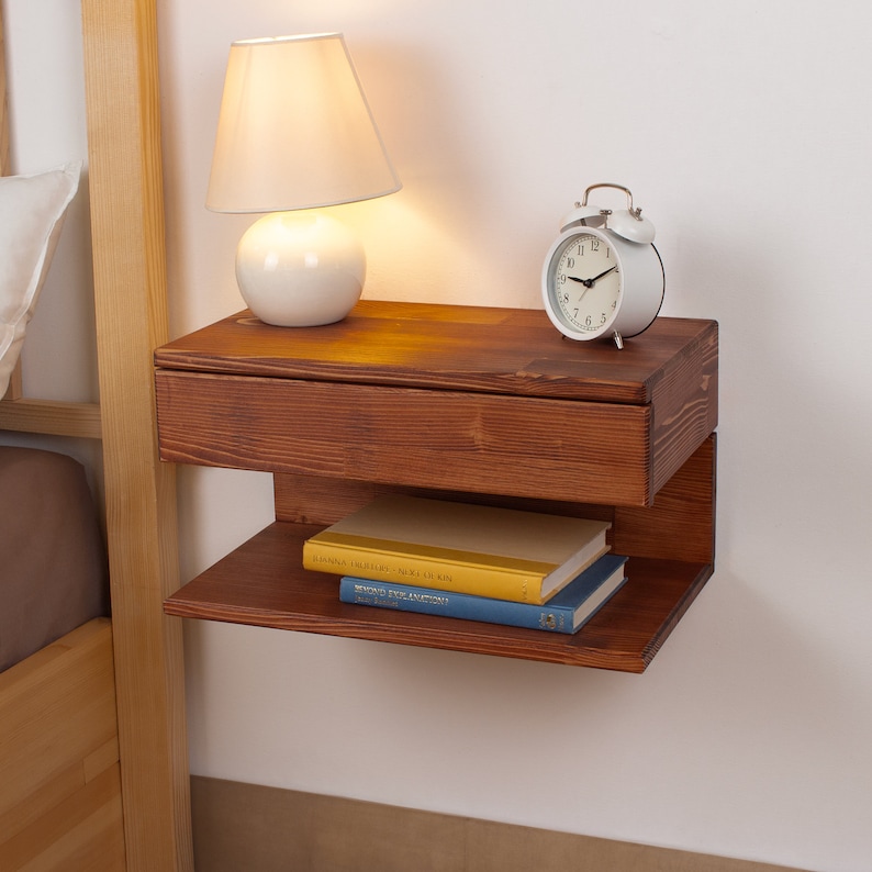 May include: A wall-mounted floating nightstand crafted from dark stained wood, featuring a single horizontal drawer at the top and an open storage shelf underneath. The unit is mounted to a wall and holds a white lamp and a white alarm clock.