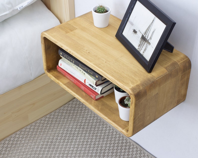 K&ouml;nnte beinhalten: Ein schwebendes Holzregal mit abgerundeten Kanten, montiert an einer wei&szlig;en Wand. Das Regal hat einen kleinen offenen Raum unter der Oberseite, in dem B&uuml;cher und eine Topfpflanze stehen. Ein gerahmtes Bild befindet sich auf der Oberseite des Regals.