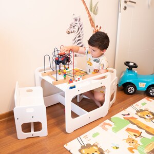 May include: A white wooden table and chair set for children, with a colorful toy on the table. The table has a built-in activity center with a bead maze and a small wooden toy truck. The chair is a small, white wooden stool. The set is on a wooden floor with a colorful play mat.