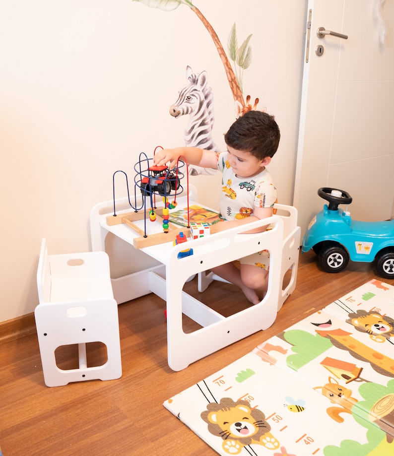 May include: A white wooden table and chair set for children, with a colorful toy on the table. The table has a built-in activity center with a bead maze and a small wooden toy truck. The chair is a small, white wooden stool. The set is on a wooden floor with a colorful play mat.
