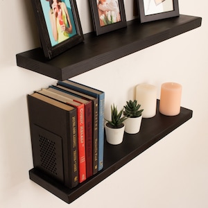 May include: Two black floating shelves with a variety of items on them, including books, candles, and plants. The shelves are mounted on a white wall.