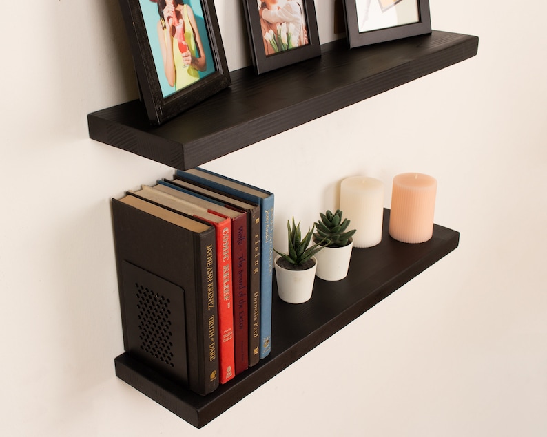 May include: Two black floating shelves with a variety of items on them, including books, candles, and plants. The shelves are mounted on a white wall.