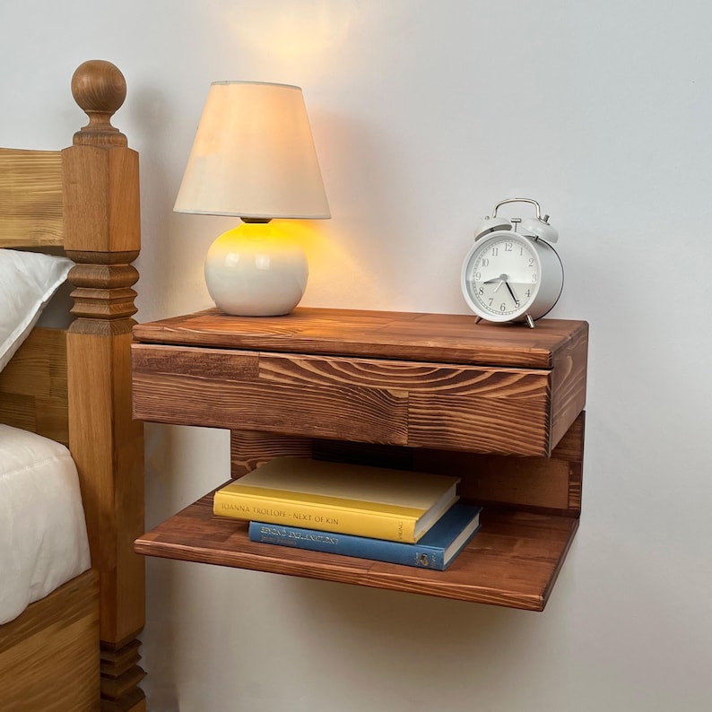 May include: A floating wooden nightstand with a dark brown wood grain finish, featuring a single drawer and an open lower shelf. A white table lamp with a glowing bulb and a silver analog alarm clock sit on top, with two books on the shelf.