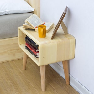 May include: A light wood nightstand with rounded edges and four legs. The nightstand has a shelf for books and a picture frame on top. A yellow mug and a book are also on top of the nightstand.