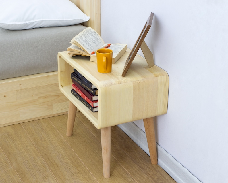 May include: A light wood nightstand with rounded edges and four legs. The nightstand has a shelf for books and a picture frame on top. A yellow mug and a book are also on top of the nightstand.