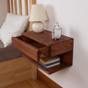May include: A wall-mounted floating nightstand crafted from dark stained wood with a visible grain pattern. The unit features a single pull-out drawer on the top section and an open storage shelf underneath, currently holding two stacked books.