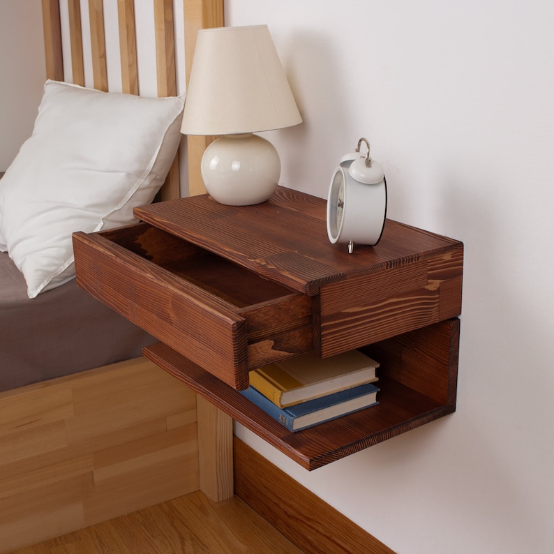 May include: A wall-mounted floating nightstand crafted from dark stained wood with a visible grain pattern. The unit features a single pull-out drawer on the top section and an open storage shelf underneath, currently holding two stacked books.