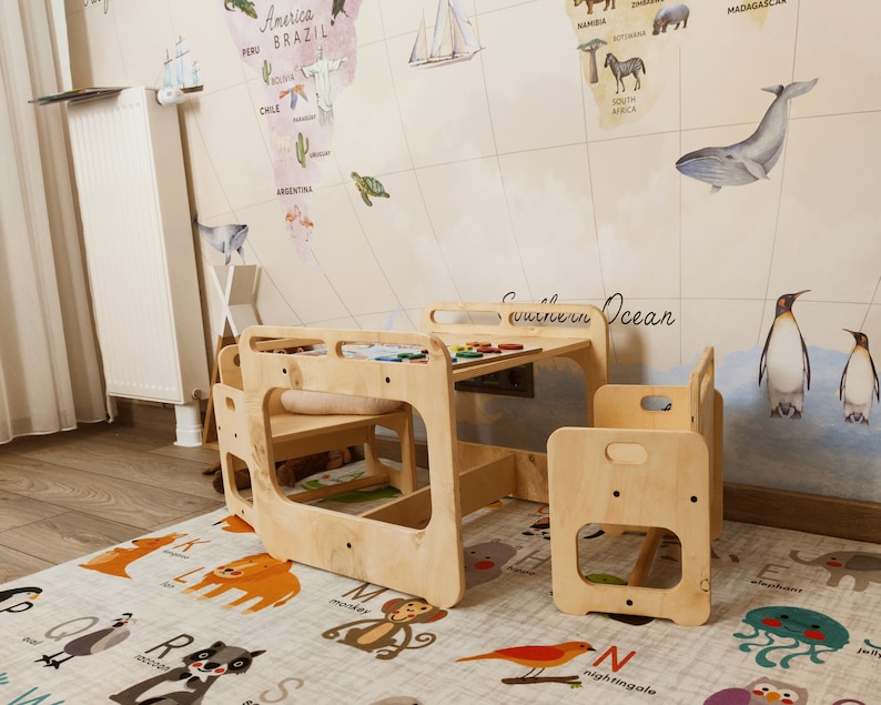 Puede incluir: Una mesa de juego de madera para ni&ntilde;os y dos sillas, colocadas sobre una alfombra con ilustraciones de animales. La mesa tiene un acabado de madera clara y un peque&ntilde;o estante debajo. Las sillas tambi&eacute;n est&aacute;n hechas de madera clara y tienen un dise&ntilde;o sencillo.