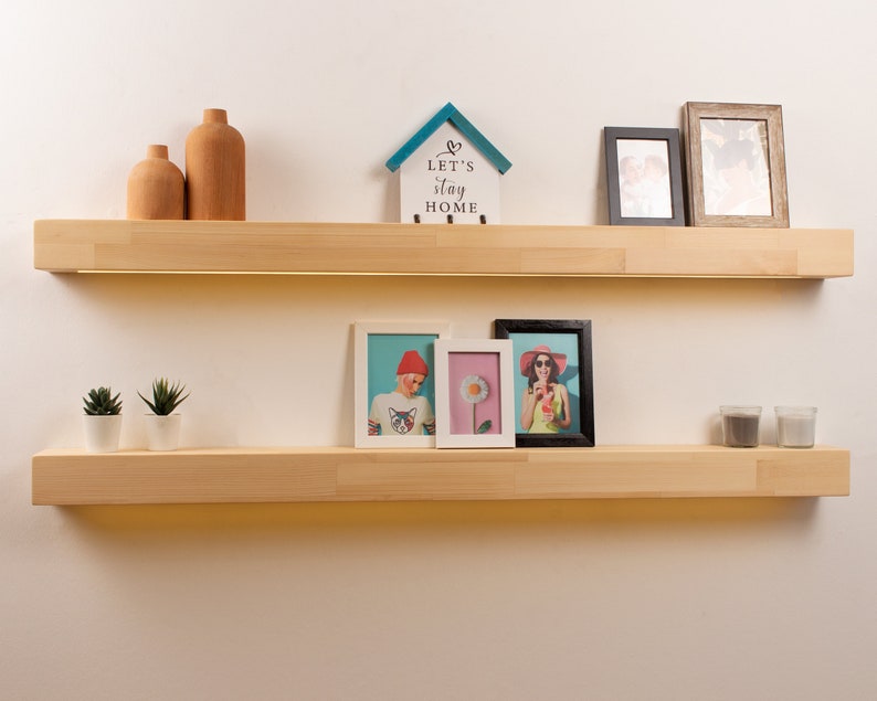 May include: Two light-colored wooden floating shelves mounted on a white wall. The top shelf holds two wooden vases, a small house-shaped sign with the text "LET'S STAY HOME", and two framed photos. The bottom shelf displays two small potted plants, and three framed photos.