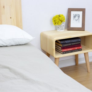 May include: A light wood nightstand with a rounded shelf and tapered legs. The nightstand has a framed photo, a vase of yellow flowers, and a stack of books on the shelf. The books include titles like "The Planet Diaries", "The Business After", "The Mystery of Things", "Comic Relief", and "Darling Ford".
