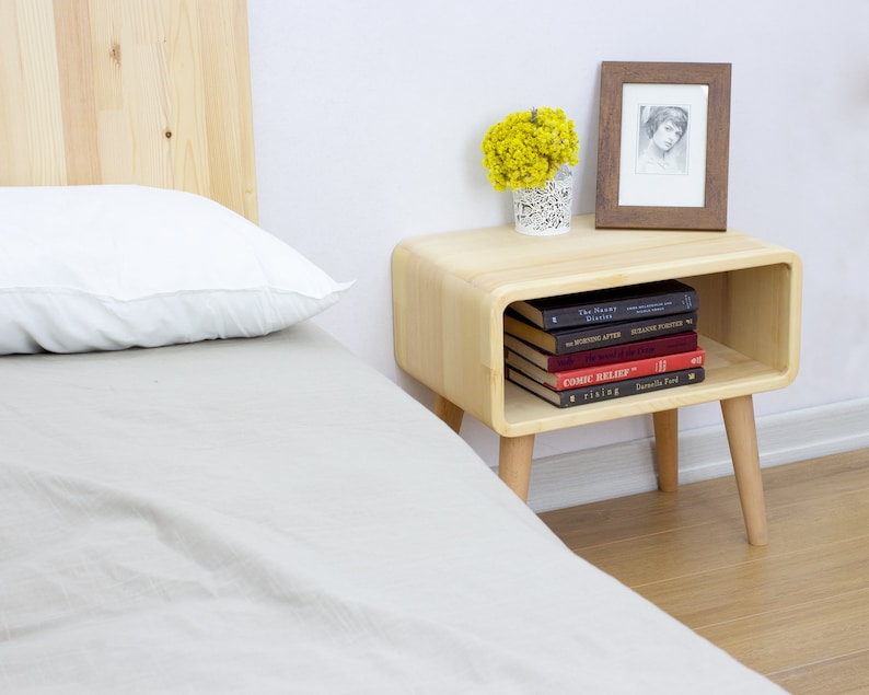 May include: A light wood nightstand with a rounded shelf and tapered legs. The nightstand has a framed photo, a vase of yellow flowers, and a stack of books on the shelf. The books include titles like "The Planet Diaries", "The Business After", "The Mystery of Things", "Comic Relief", and "Darling Ford".