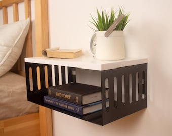 May include: A white and black floating shelf with a black metal frame and a white top. The shelf is mounted on a wall and has two books on it. The books are titled "The Glory" and "The Flaming Rising". A potted plant with green leaves is on the shelf.
