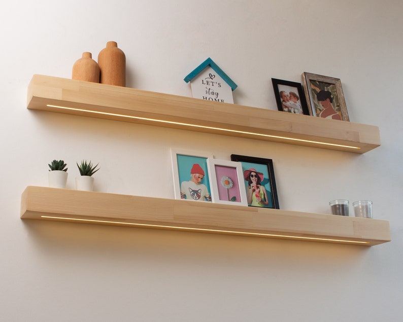 May include: Two light-colored wooden floating shelves with built-in LED lighting. The shelves are mounted on a white wall and display decorative items such as small plants, framed photos, and wooden vases. The shelves are approximately 48 inches long.