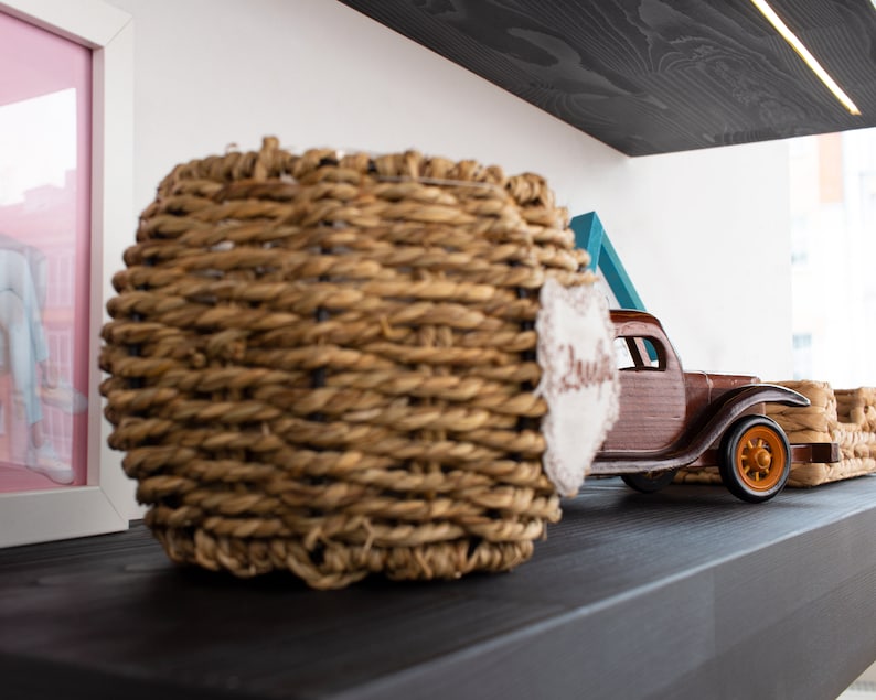 K&ouml;nnte beinhalten: Ein geflochtener Korb mit einem Etikett mit der Aufschrift "Sweet" neben einem Holzspielzeug-LKW mit orangefarbenen R&auml;dern. Der Korb besteht aus einem hellbraunen Material und ist rund. Der LKW ist braun und hat ein detailliertes Design.
