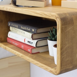 K&ouml;nnte beinhalten: Ein schwebendes Holzregal mit abgerundeter Kante, auf dem ein Stapel B&uuml;cher und zwei Topfpflanzen stehen. Das Regal ist an einer Wand montiert und hat eine nat&uuml;rliche Holzoptik.