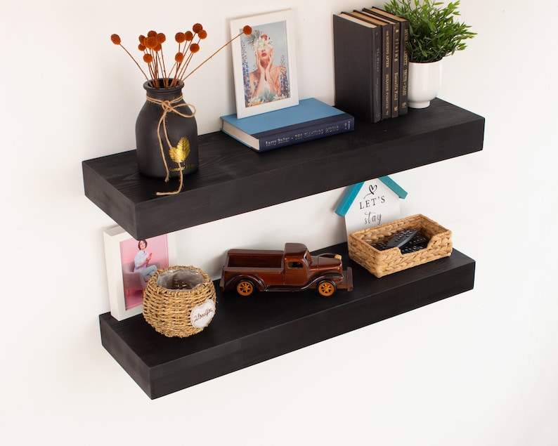K&ouml;nnte beinhalten: Zwei schwarze schwebende Regale an einer wei&szlig;en Wand, jeweils mit dekorativen Gegenst&auml;nden. Das obere Regal zeigt eine Vase mit getrockneten Blumen, ein gerahmtes Bild und B&uuml;cher. Das untere Regal zeigt einen Spielzeug-LKW, ein gerahmtes Bild und einen Weidenkorb.