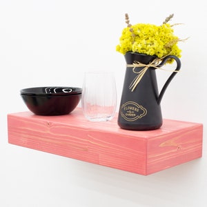 May include: A pink wooden shelf with a black pitcher filled with yellow flowers, a black bowl, and a clear glass. The pitcher has the text "Flowers & Garden" on it.