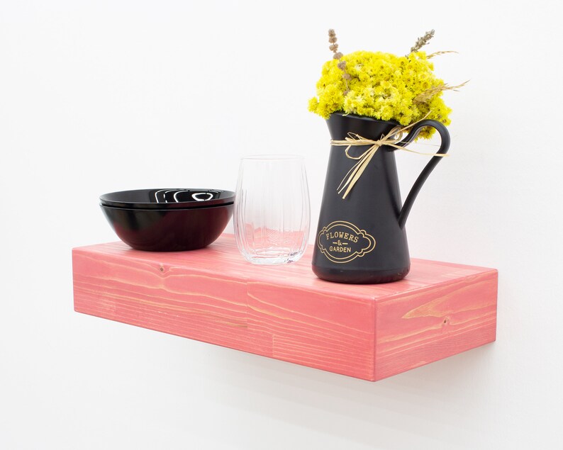 May include: A pink wooden shelf with a black pitcher filled with yellow flowers, a black bowl, and a clear glass. The pitcher has the text "Flowers & Garden" on it.