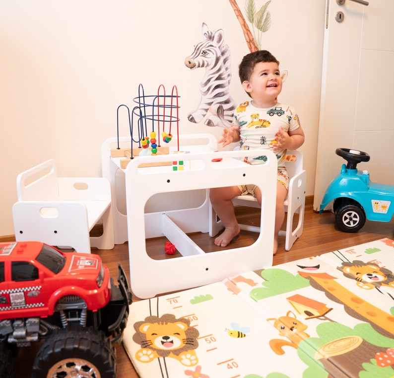 May include: A white wooden play table and chairs set with a colorful bead maze toy. A child sits at the table, and a red toy truck and a blue toy car are on the floor. A colorful play mat with animal designs is on the floor.