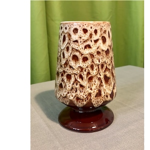 Może przedstawiać: Wazon z ceramiki w stylu vintage o teksturowanej, wzorzystej powierzchni. Wazon ma brązowo-kremową kolorystykę z rozszerzaną górą i okrągłą, ciemnobrązową podstawą. Wzór ma strukturę plastra miodu.