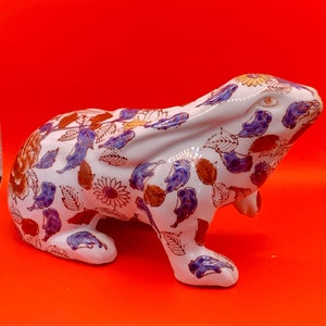 May include: A ceramic rabbit figurine with a white base, adorned with a floral pattern in shades of blue, brown, and gold. The rabbit is posed in a seated position, with its ears perked up. The figurine is set against a vibrant red background.