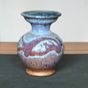 Puede incluir: Un jarrón de cerámica con cuerpo bulboso y borde acampanado. El jarrón presenta un esmalte moteado azul y granate. La base es de color marrón claro. El jarrón se exhibe sobre una superficie de madera.